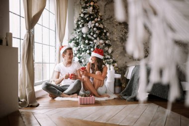 Beyaz tişörtler ve Noel Baba şapkalı adam ve kız, yeni yıl ağacının yanındaki pencerenin önünde kırmızı bardaklarla oturuyorlar, hediyeler ve mumlar