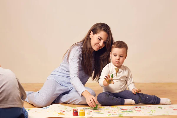 Piękny szczęśliwy młoda matka i jej synka, ubrany w domu ubrań siedzą na drewnianej podłodze w pokojach i malowanie palcami na białym papierze — Zdjęcie stockowe