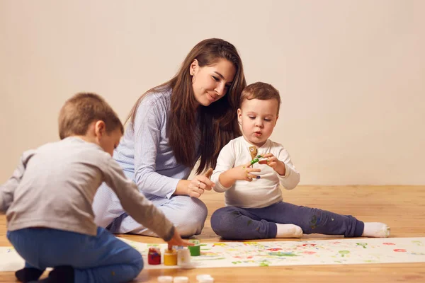 Щаслива молода мама і двоє її маленьких синів, одягнені в домашній одяг, сидять на дерев'яній підлозі в кімнаті і малюють пальцями на білому папері — стокове фото