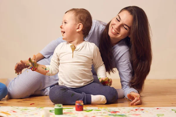Szczęśliwą matką i jej synka z farby na jego twarzy, ubranego w domu ubrań siedzą na drewnianej podłodze w pokojach i malowanie palcami na białym papierze — Zdjęcie stockowe
