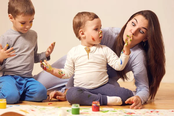 Szép boldog anya és két kis fia öltözött otthoni ruhát ül a fa padló a szobában, és a festészet ujjaival a fehér könyvről — Stock Fotó
