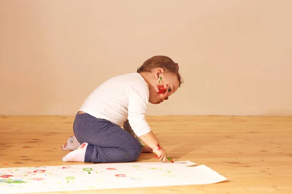 Μικρό αγόρι ντυμένος στο σπίτι ρούχα κάθονται στο ξύλινο πάτωμα στο δωμάτιο και ζωγραφική με τα δάχτυλα στο χαρτί — Φωτογραφία Αρχείου
