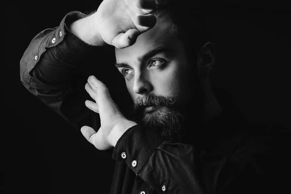 Retrato preto e branco de um homem elegante com uma barba e penteado elegante vestido com a camisa preta segurando as mãos na frente de seu rosto no fundo preto — Fotografia de Stock