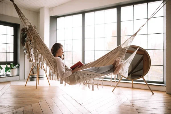 Fille aux cheveux foncés vêtue de pantalon, pull et pantoufles chaudes lit un livre couché dans un hamac dans une chambre confortable avec sol en bois et fenêtres panoramiques et un miroir rond sur le sol — Photo