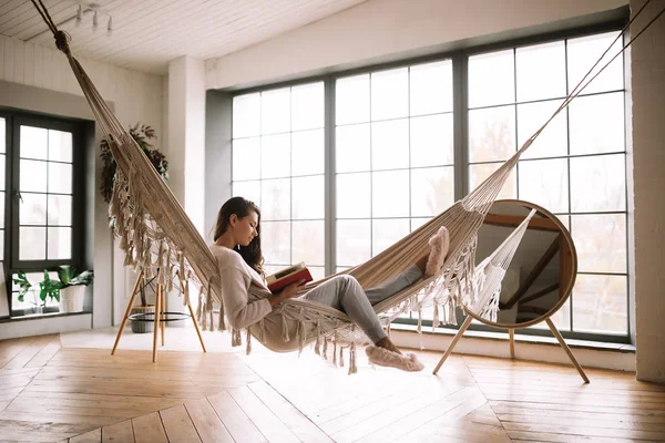 Fille aux cheveux foncés vêtue de pantalon, pull et pantoufles chaudes lit un livre couché dans un hamac dans une chambre confortable avec sol en bois et fenêtres panoramiques et un miroir rond sur le sol — Photo