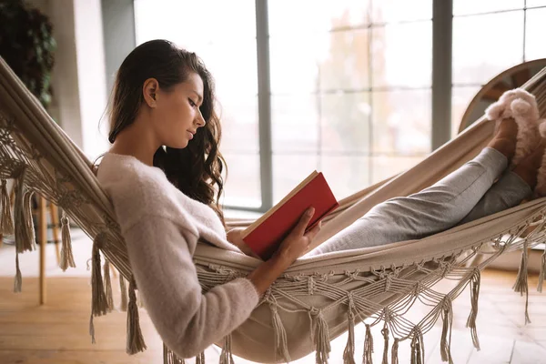 Dark-haired girl dressed in pants, sweater and warm slippers reads a book lying in a hammock in a cozy room with panoramic windows — Stock Photo, Image