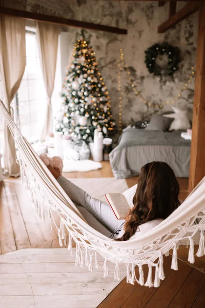 Красивая темноволосая девушка в брюках, свитере и теплых тапочках читает книгу, лежащую в гамаке в уютной украшенной комнате с новогодней елкой — стоковое фото