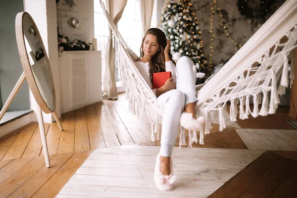 Schönes dunkelhaariges Mädchen in Hose, Pullover und warmen Hausschuhen liest ein Buch, das in einer Hängematte in einem gemütlich dekorierten Raum mit einem Neujahrsbaum liegt — Stockfoto