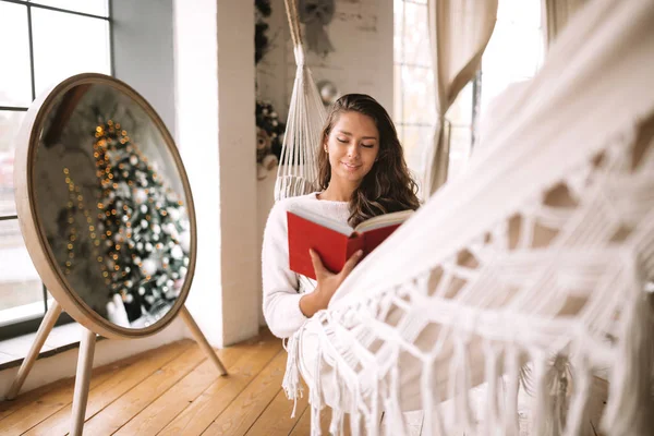 Красивая темноволосая девушка в штанах и свитере читает книгу, лежащую в гамаке рядом с круглым зеркалом на полу в уютной комнате — стоковое фото