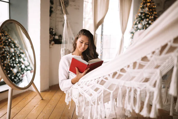 Belle fille aux cheveux bruns habillée en pantalon et pull lit un livre couché dans un hamac à côté du miroir rond sur le plancher dans une chambre décorée confortable avec un arbre du Nouvel An — Photo