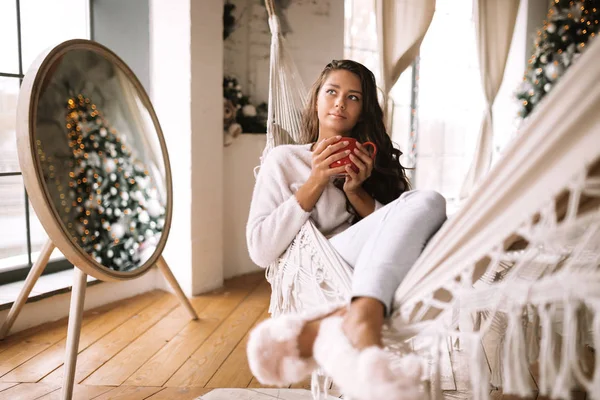 Charmant donkerharige meisje gekleed in broek, trui en warme pantoffels houdt een rode kop liggend in een hangmat in een gezellig ingerichte kamer met een nieuwjaarsboom — Stockfoto