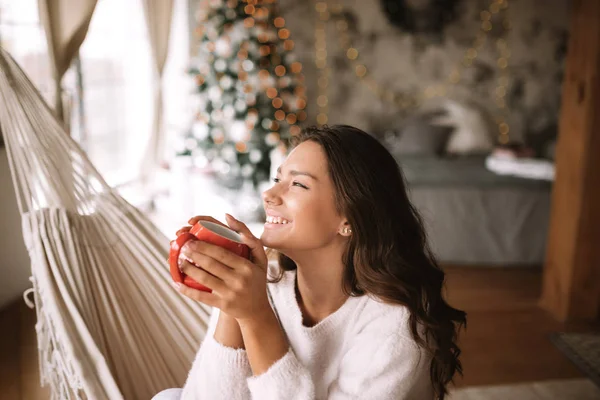 Усміхнена темноволоса дівчина, одягнена в бежевий светр і штани, тримає червону чашку, сидячи в гамаку в затишній прикрашеній кімнаті з новорічною ялинкою — стокове фото