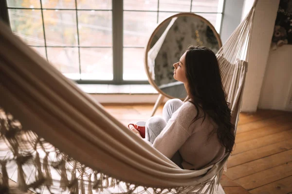 Visão traseira de uma menina sentada em uma rede em um quarto acolhedor com piso de madeira e janelas panorâmicas e um espelho redondo no chão — Fotografia de Stock