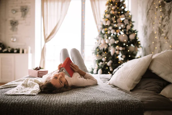 Очаровательная девушка в белом свитере и брюках читает книгу, лежащую на кровати с серым одеялом, белыми подушками и новогодним подарком в украшенной комнате с новогодней елкой — стоковое фото