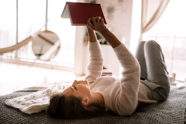 Charmante fille vêtue d'un pull et d'un pantalon blancs lit un livre couché sur le lit avec une couverture grise, des oreillers blancs et un cadeau de Nouvel An dans une salle de cory — Photo