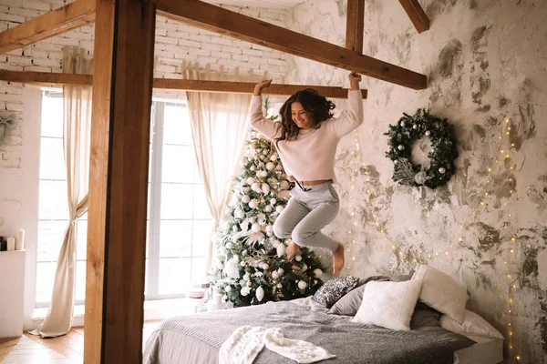 Charmant meisje gekleed in witte trui en broek springt op het bed met grijze deken en witte kussens in een gezellig ingerichte kamer met een nieuwe jaar boom — Stockfoto