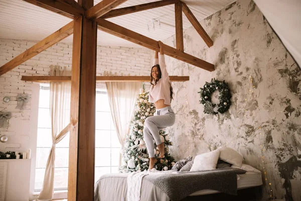 Linda chica vestida con suéter blanco y pantalones está colgando en una barra de madera por encima de la cama con manta gris y almohadas blancas en una acogedora habitación decorada con un árbol de Año Nuevo — Foto de Stock