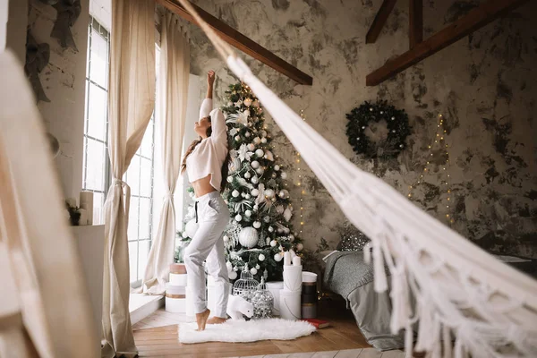 Charming girl dressed in white sweater and pants stands next to the New Year tree in front of the window and stretches up  in a cozy decorated room  with New Years wreath — Stock Photo, Image