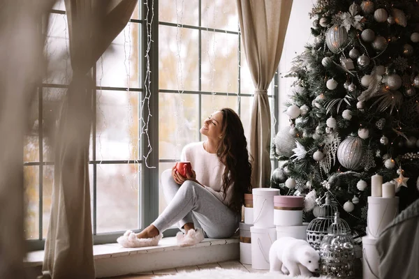 Bájos sötét hajú lány öltözött nadrágot, pulóvert és meleg papucs tart egy piros csésze ül az ablakpárkányon egy panoráma ablak a szobában mellett az új év fa, ajándékok és gyertyák — Stock Fotó