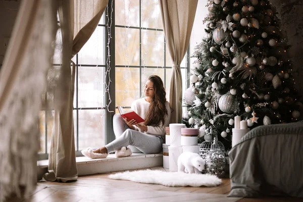 Nice dark-haired girl dressed in pants, sweater and warm slippers reads a book sitting on the windowsill of a panoramic window in the room next to the New Year tree, gifts and candles