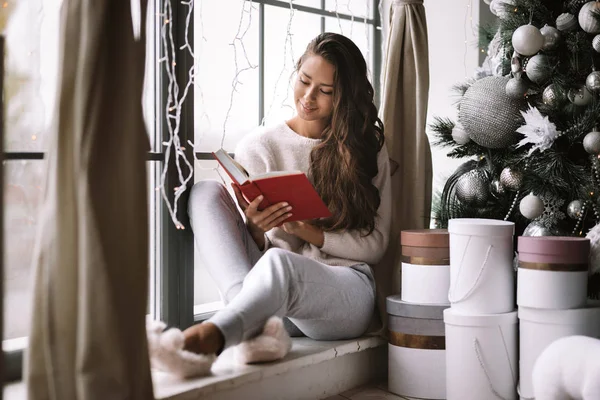 Гарна темноволоса дівчина, одягнена в штани, светр і теплі тапочки читає книгу, що сидить на підвіконні панорамного вікна в кімнаті поруч з новорічною ялинкою, подарунками і свічками — стокове фото