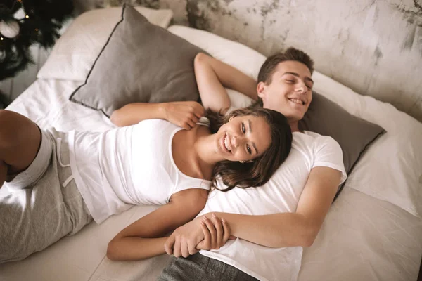 Glückliche Kerle und Mädchen in weißen T-Shirts liegen auf einem Bett mit einer weißen Decke mit grauen Kissen und halten sich an den Händen — Stockfoto