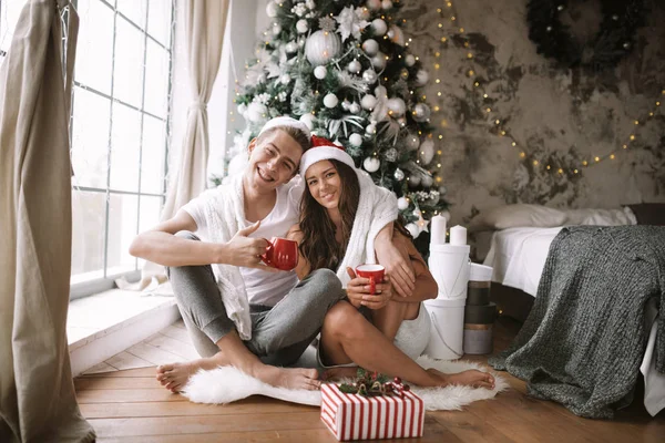 Ευτυχισμένο αγόρι και κορίτσι με λευκά μπλουζάκια και τα καπέλα του Άη Βασίλη κάθονται με κόκκινες κούπες στο πάτωμα μπροστά από το παράθυρο δίπλα στο δέντρο της Πρωτοχρονιάς, δώρα και κεριά — Φωτογραφία Αρχείου