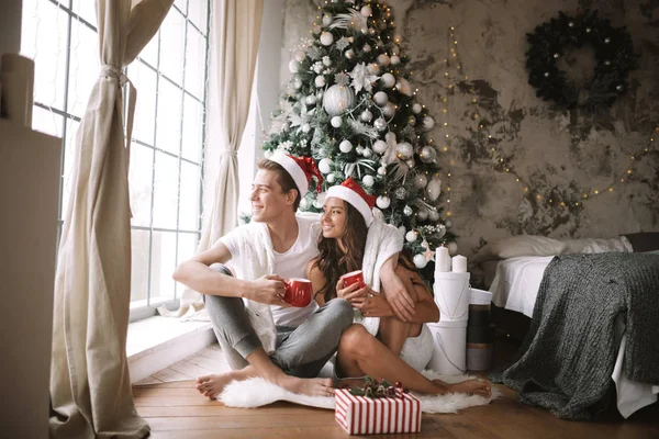 Щасливий хлопець і дівчина в білих футболках і Дід Мороз шапки сидять з червоними чашками на підлозі перед вікном поруч з новорічним деревом, подарунками і свічками — стокове фото