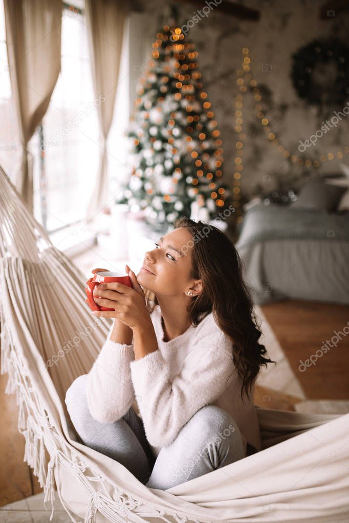 Charming dark-haired girl dressed in beige sweater and pants holds a red cup sitting in a hammock in a cozy decorated room with a New Year tree