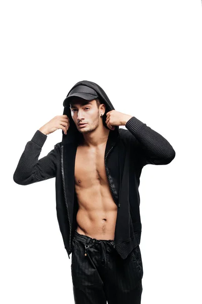 Bonito jovem dançarino vestido com uma camisola em um tronco nu com um capuz no boné e calças pretas fica com as mãos no capô — Fotografia de Stock