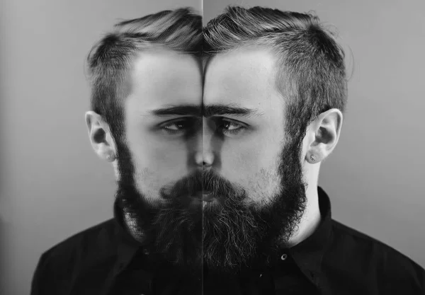 Foto en blanco y negro de un hombre con barba y peinado elegante vestido con la camisa negra de pie junto al espejo con reflejo de parte de su rostro — Foto de Stock