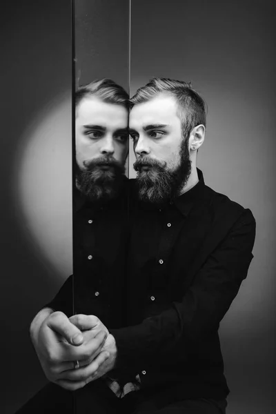 Foto en blanco y negro de un hombre con barba y peinado elegante vestido con la camisa negra de pie junto al espejo con su reflejo — Foto de Stock