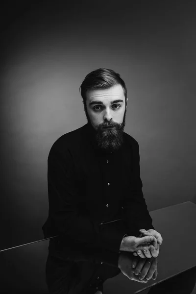 Foto en blanco y negro de un hombre con barba y peinado elegante vestido con la camisa negra de pie junto al espejo con las manos sobre ella — Foto de Stock