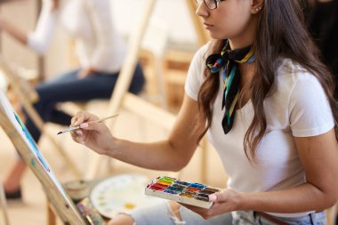 Boynunda bir eşarp ile beyaz t-shirt ve kot giymiş gözlük kahverengi saçlı kız sanat stüdyosunda bir resim boyar