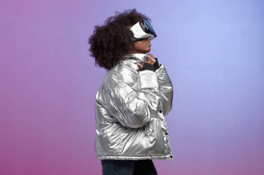 Trendy kıvırcık kahverengi saçlı kız gümüş renkli ceket giymiş neon arka plan üzerinde stüdyoda sanal gerçeklik gözlük kullanır