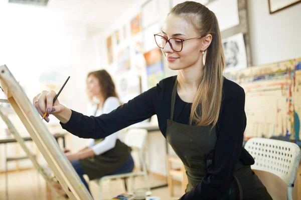 Krásná světlovlasá dívka v brýlích oblečených v černé blůze a zástěra sedí na stojanu a maluje obraz v uměleckým studiu — Stock fotografie