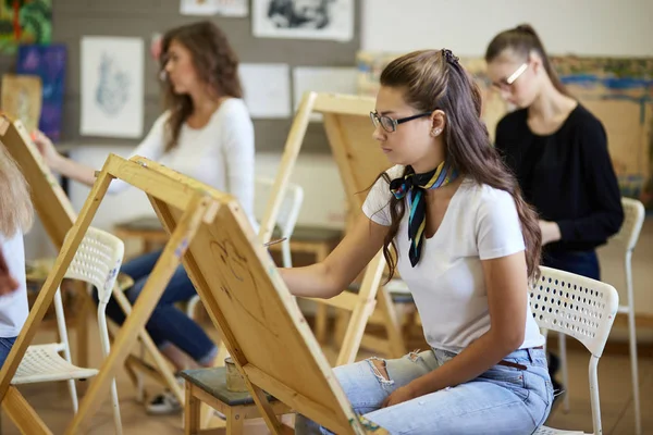 Lección de dibujo en el estudio de arte. Árbol encantadoras chicas jóvenes pintar imágenes sentadas en los caballetes — Foto de Stock