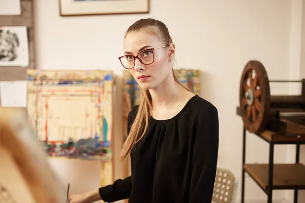 Sjarmerende lyshåret jente i briller kledd i svart bluse setter seg ved staffeliet og maler et bilde i kunststudioet – stockfoto