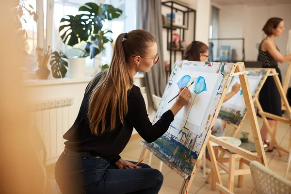 Encantadora chica de pelo claro en gafas vestidas con blusa negra y jeans se sienta en el caballete y pinta un cuadro en el estudio de arte — Foto de Stock