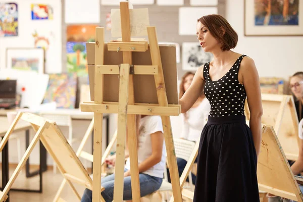 Professor de desenho encantador no vestido bonito mostra técnica de desenho no cavalete no estúdio de arte — Fotografia de Stock