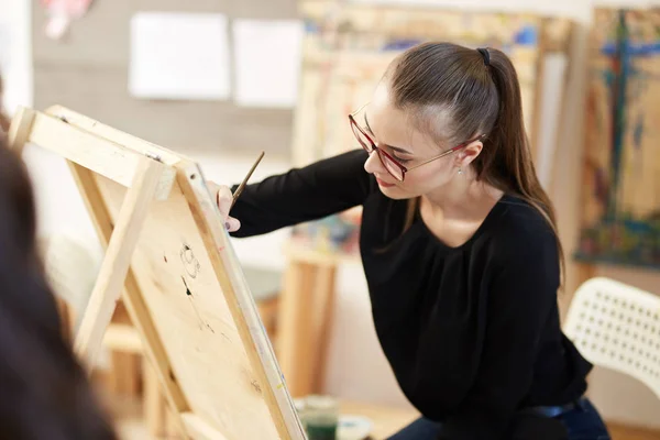 Krásná světlovlasá dívka v brýlích oblečených v černé blůze sedí na stojanu a maluje obraz v uměleckým studiu — Stock fotografie