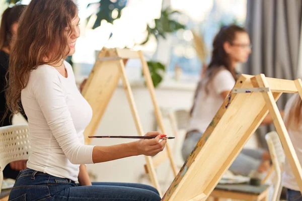 Menina sorridente com cabelo encaracolado marrom vestido com blusa branca pinta um quadro no cavalete na escola de desenho — Fotografia de Stock