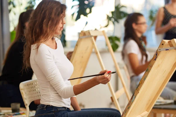 Tânără fată fermecătoare cu păr creț maro îmbrăcat în bluză albă pictează o imagine la șevalău în școala de desen — Fotografie, imagine de stoc