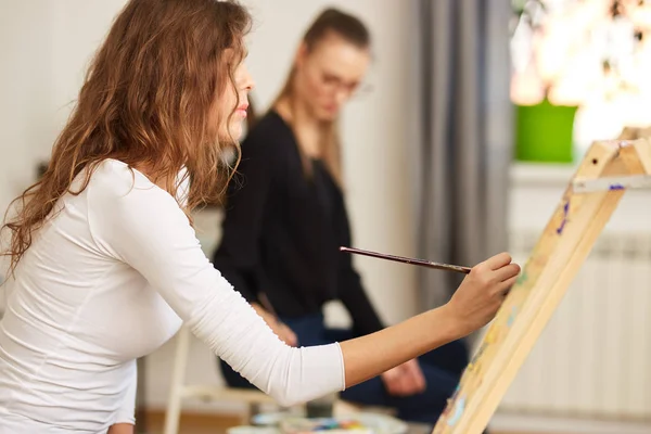 Menina com cabelo encaracolado marrom vestido com blusa branca pinta um quadro no cavalete na escola de desenho — Fotografia de Stock