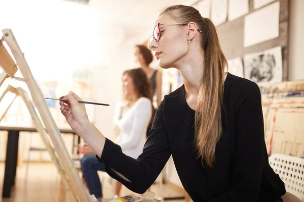 Niza joven en gafas vestidas con blusa negra y jeans se sienta en el caballete y pinta un cuadro en la escuela de dibujo — Foto de Stock