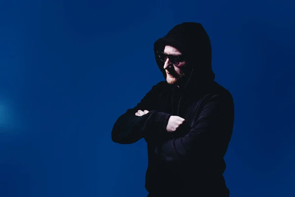 Ritratto di uomo di moda in maglione nero con cappuccio e occhiali da sole in luce al neon in studio — Foto Stock