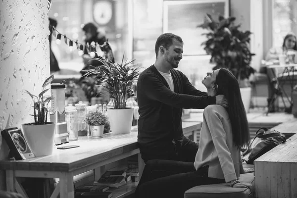 Glücklicher liebevoller Kerl legt seine Hände auf die Schultern der Mädchen, die am Tisch im Café sitzen und sie anschauen. Schwarz-Weiß-Foto — Stockfoto