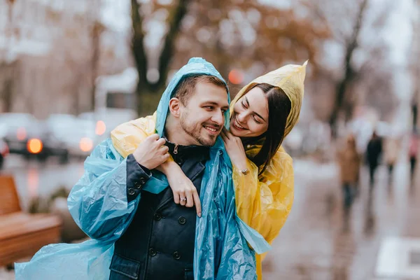 Happy couple loving guy and his girlfriend dressed in raincoats are hugging on the street in the rain