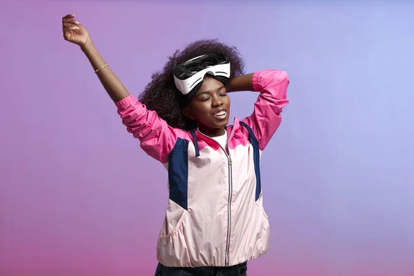 Funny curly brown-haired girl dressed in the pink sports jacket is wearing on her head the virtual reality glasses in the studio on neon background — Stock Photo, Image