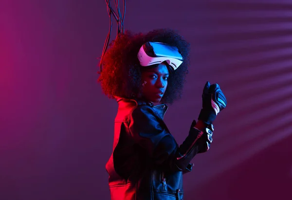 Lockiges dunkelhaariges Mädchen in schwarzer Lederjacke und Handschuhen trägt die Virtual-Reality-Brille auf dem Kopf im dunklen Studio mit Neonlicht — Stockfoto